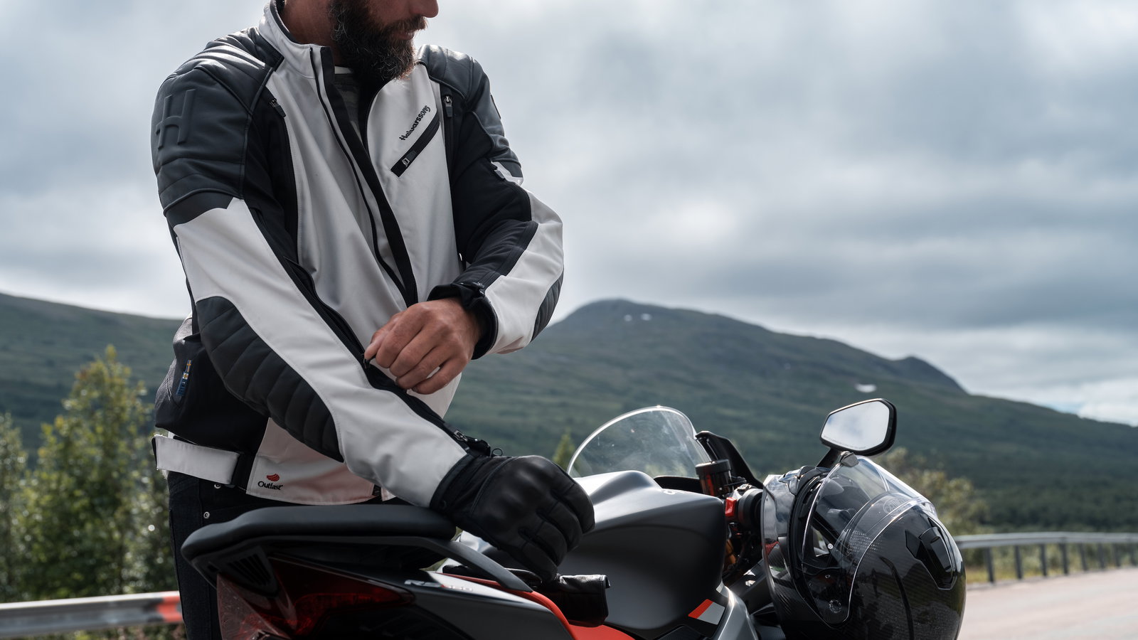 HALVARSSONS SOLBERG Person with beard standing over a motorcycle. White jacket on show. Mountains in background.